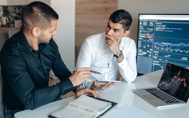 Traders in conversation in front of multi-monitor workstations displaying financial charts